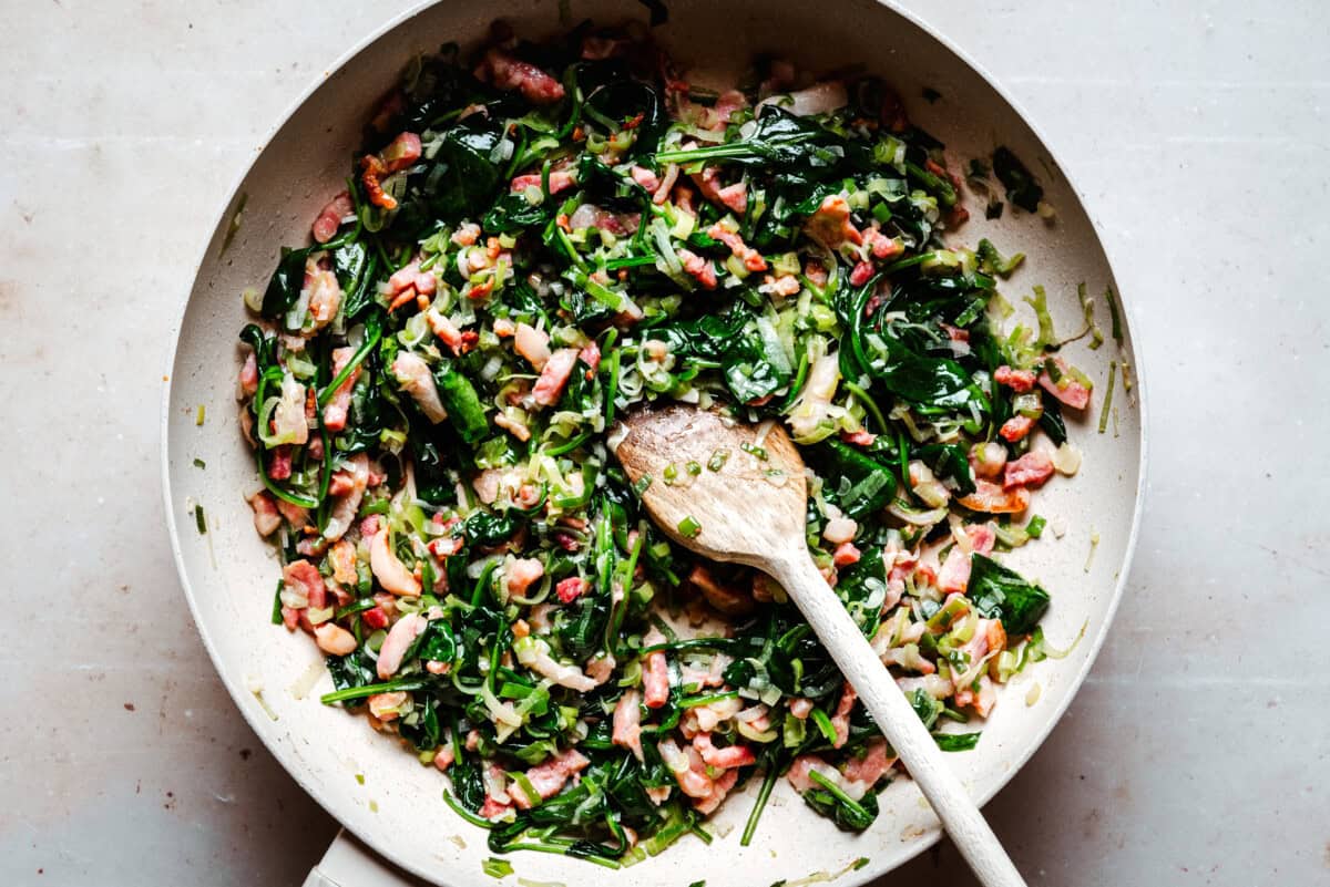 A frying pan filled with sautéed spinach, bacon, onions, and herbs sits on a light surface, with a wooden spoon resting in the mixture.