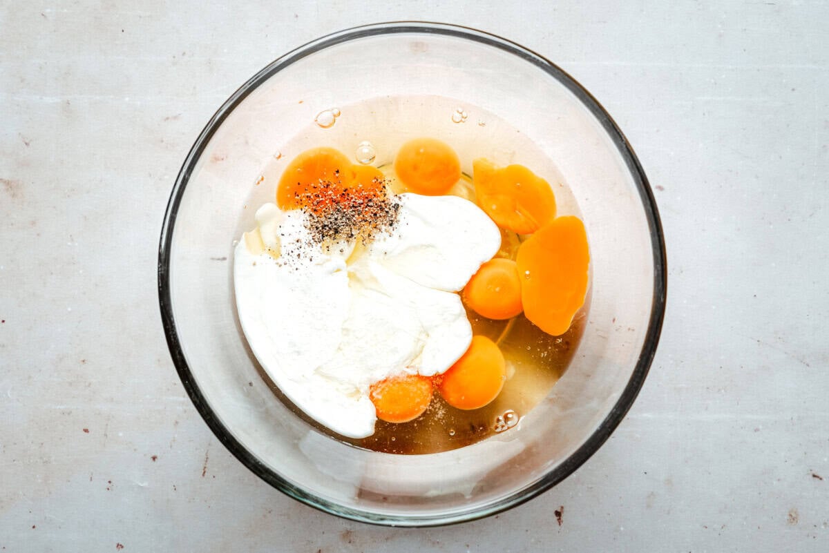 A glass bowl containing cream, eggs, oil, salt, and pepper on a light-colored surface, seen from above.