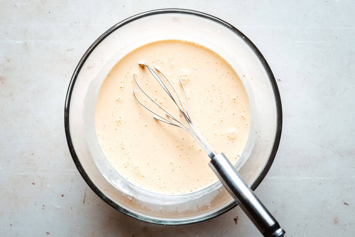A metal whisk resting in a glass bowl filled with custard on a light-colored surface.