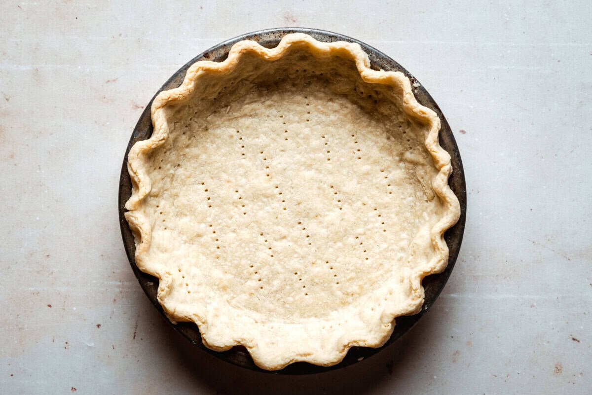A baked, empty pie crust with crimped edges sits in a metal pie pan. The crust surface has small fork holes evenly spaced across the bottom. The background is a light, neutral countertop.