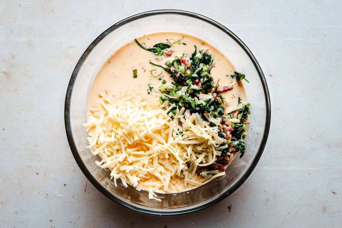 A glass bowl contains shredded cheese, a mixture of sautéed spinach and bacon, and a creamy custard mixture, arranged side by side on a light countertop.