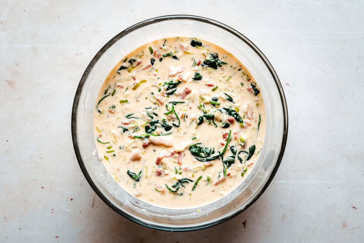 A glass bowl filled with a creamy mixture containing spinach, bacon, and herbs sits on a light-colored surface.