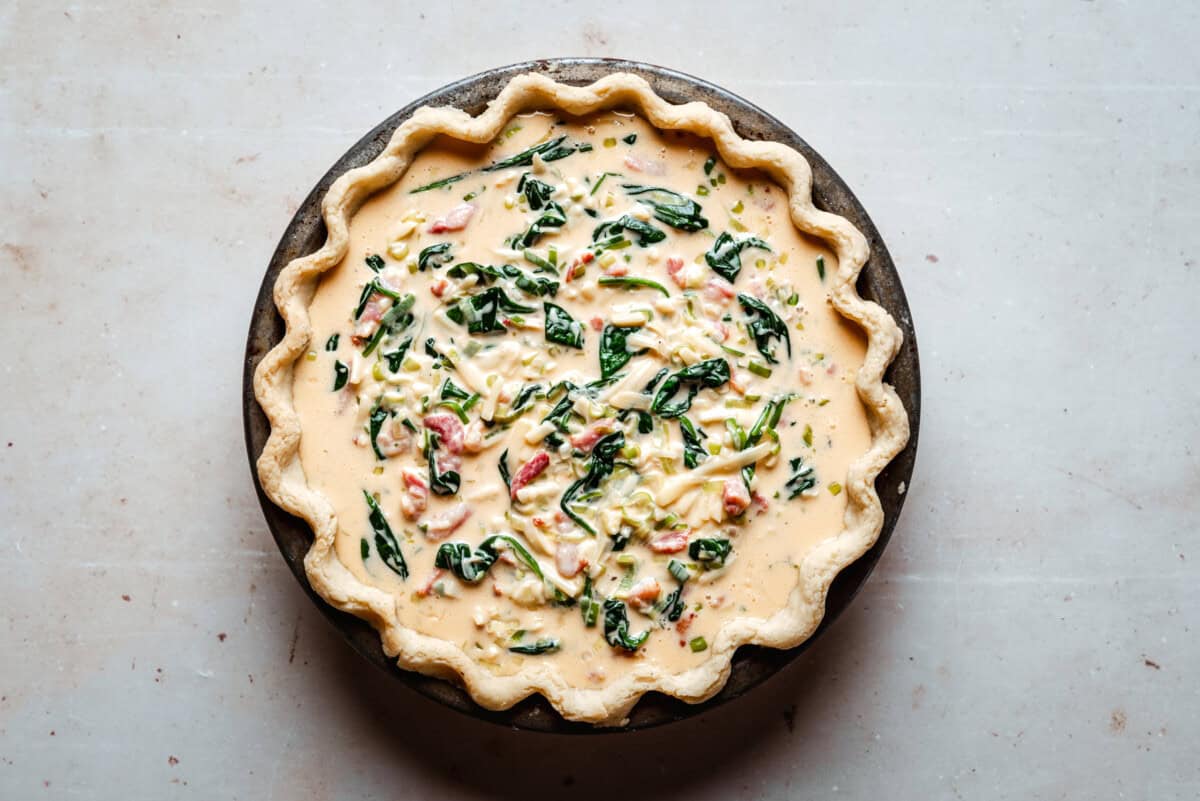 Unbaked quiche with a fluted pie crust filled with a creamy egg mixture, spinach, and diced vegetables, placed on a light-colored surface.