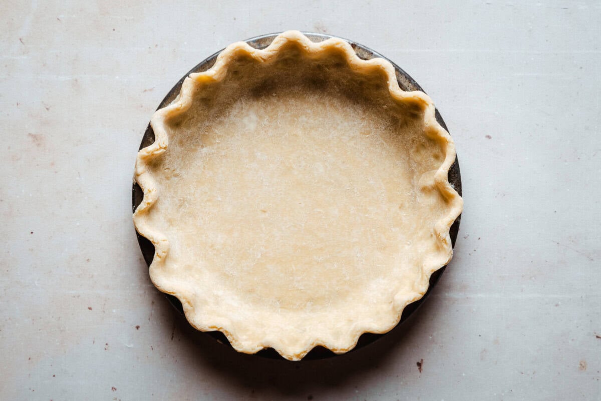 A round, unbaked pie crust with fluted edges sits in a metal pie pan on a light-colored surface.