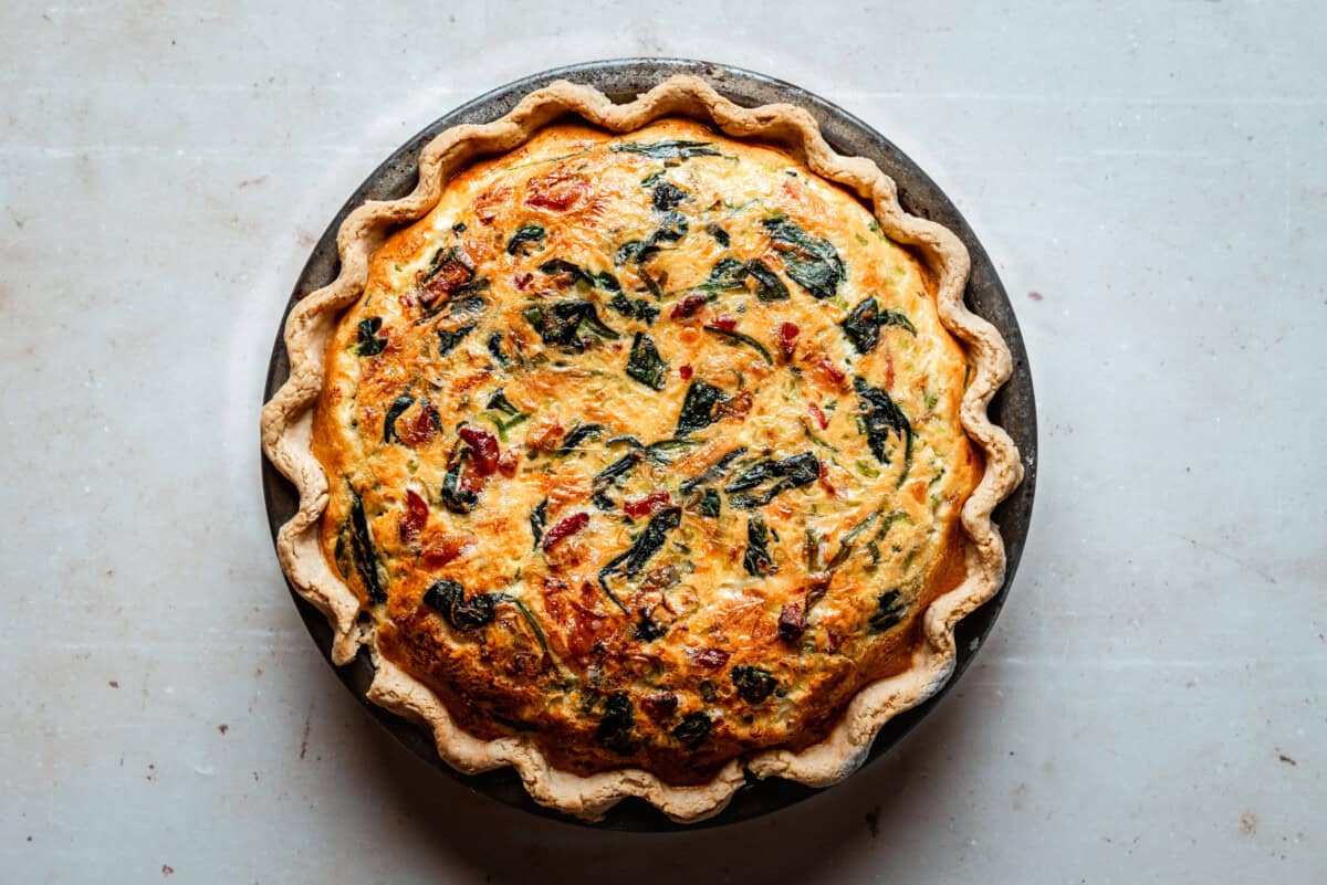 A freshly baked spinach bacon quiche with a golden, flaky crust, filled with visible leafy greens and bits of red and brown ingredients, sits on a round metal pie pan against a light-colored surface.