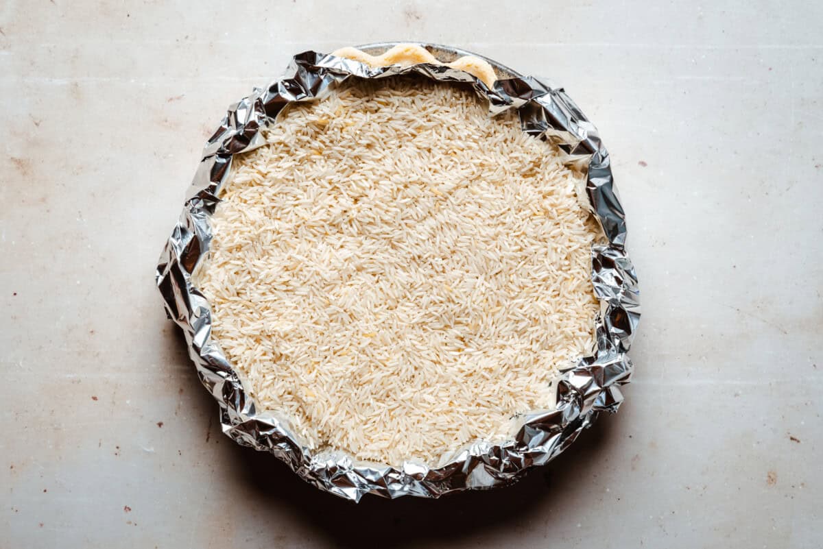 Unbaked pie crust round metal pie tin lined with foil and filled with uncooked rice, set on a light-colored countertop. The rice is being used as a pie weight for blind baking.