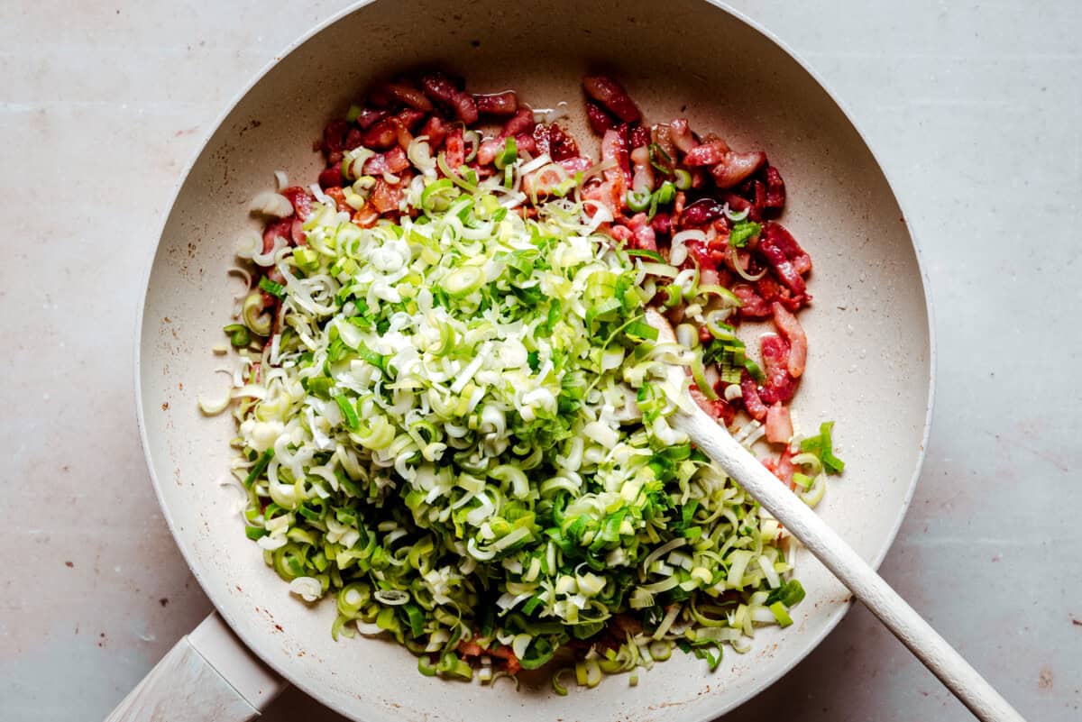 A pan filled with chopped leeks and pieces of bacon being stirred with a wooden spoon on a light surface.
