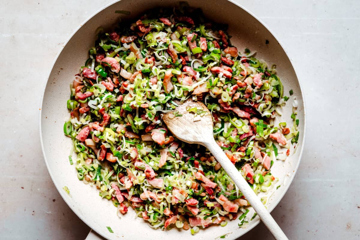 A sauté pan filled with chopped leeks, bacon, and herbs being stirred with a wooden spoon on a light countertop.