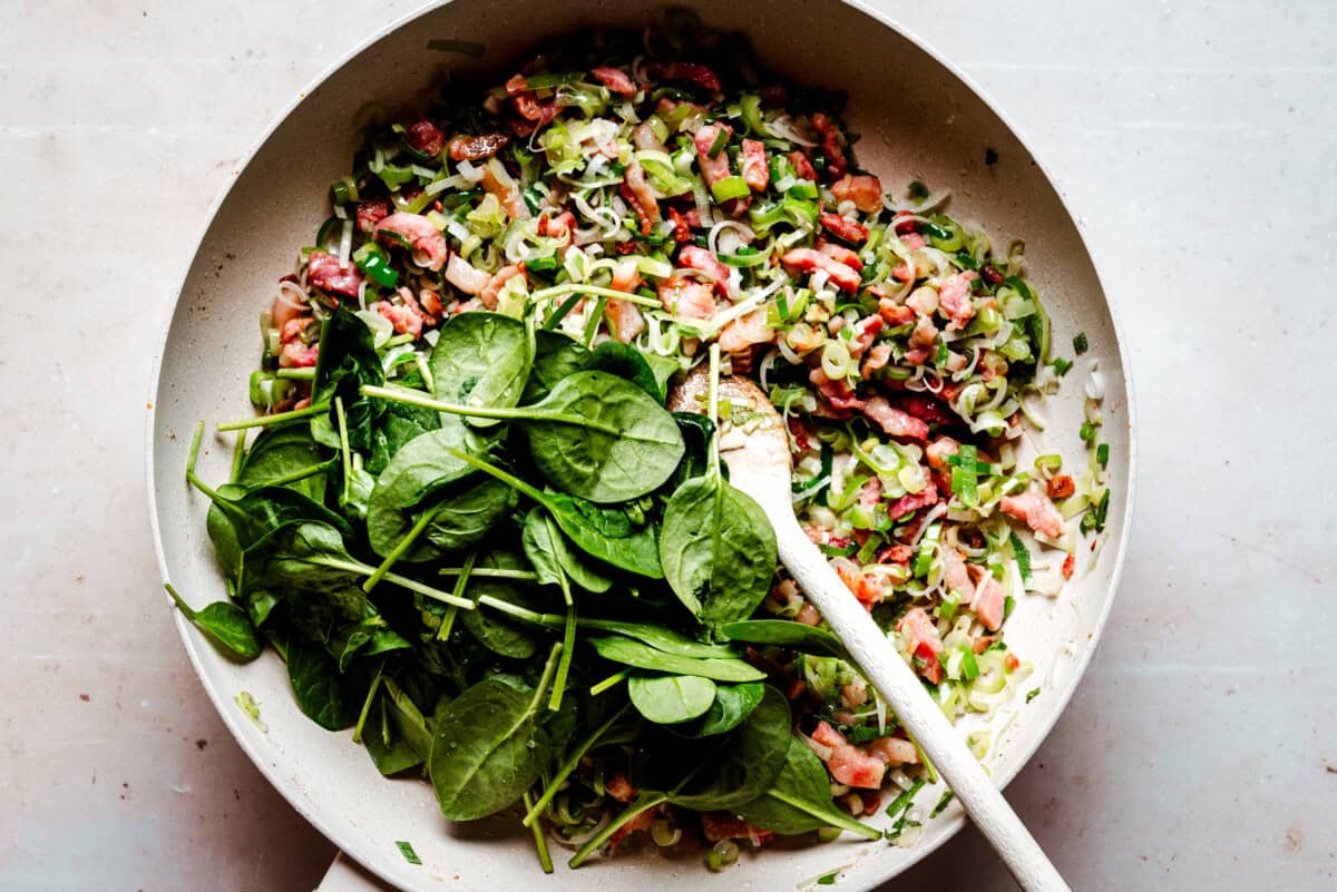 A skillet with chopped bacon, green onions, and fresh spinach being stirred by a wooden spoon on a light countertop.