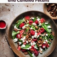 A bowl of spinach salad with feta cheese, candied pecans, fresh raspberries, and pink dressing on a textured surface, with extra pecans and dressing on the side. Text above promotes spinach salad with feta and candied pecans.