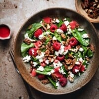 A bowl of spinach salad topped with raspberries, candied pecans, and crumbled feta cheese, with pink raspberry vinaigrette drizzled on top and extra dressing in a small cup nearby.
