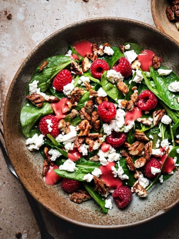 A bowl of spinach salad topped with raspberries, candied pecans, and crumbled feta cheese, with pink raspberry vinaigrette drizzled on top and extra dressing in a small cup nearby.
