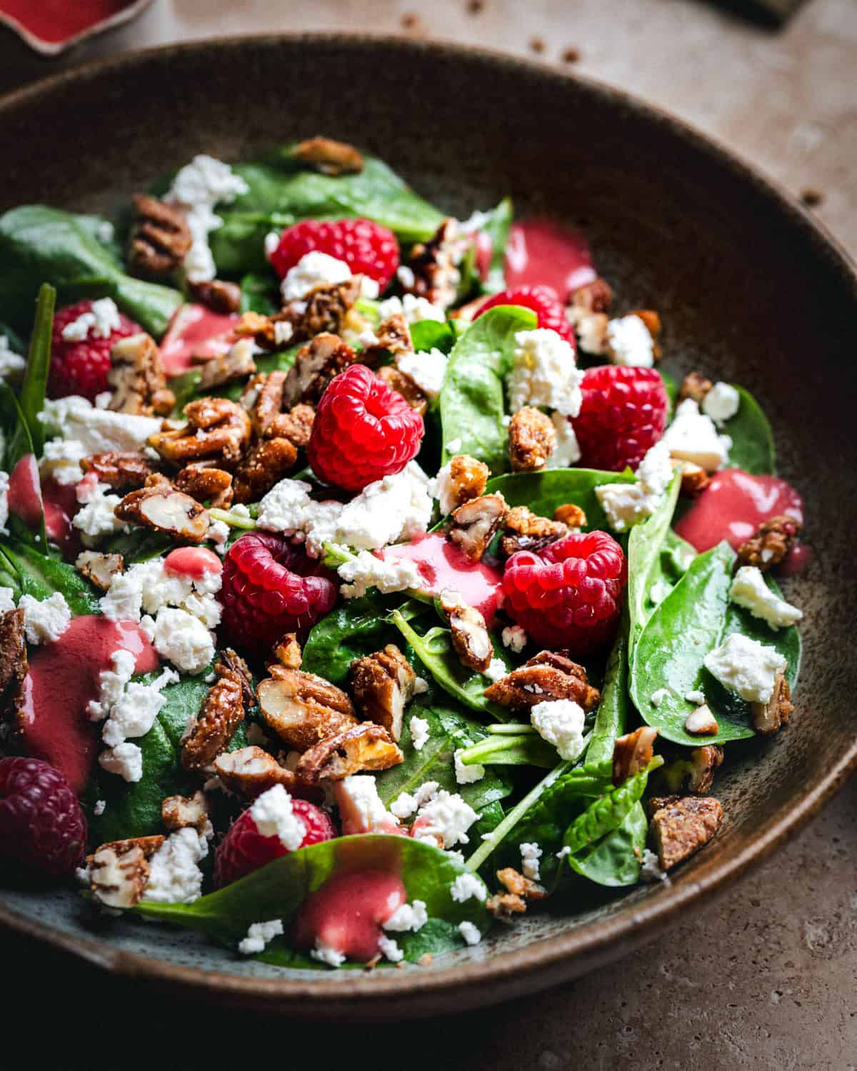 A bowl of fresh spinach salad topped with raspberries, crumbled gorgonzola cheese, candied pecans, and drizzled with a pink raspberry vinaigrette dressing.