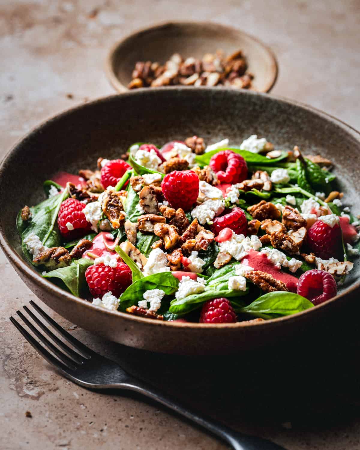 A bowl of fresh salad with spinach leaves, raspberries, crumbled gorgonzola cheese, and candied pecans, with a small dish of pecans and a fork beside the bowl on a rustic surface.