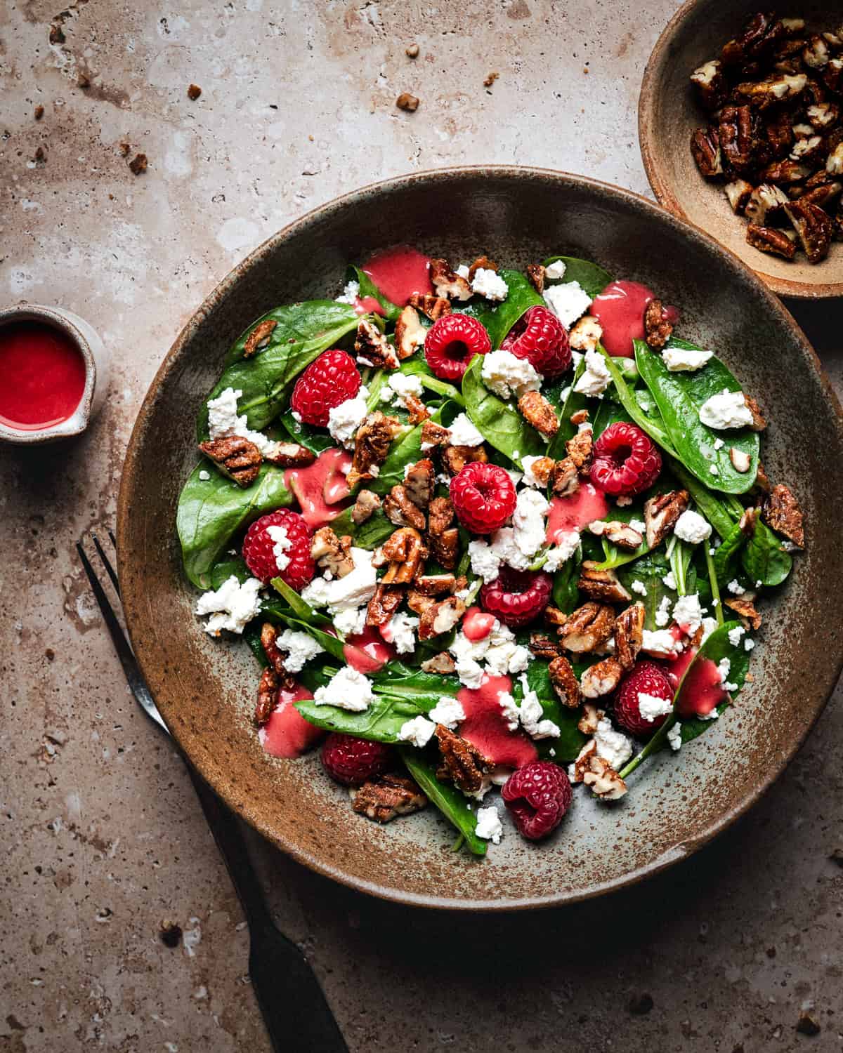 A bowl of spinach salad with raspberries, crumbled gorgonzola, candied pecans, and raspberry vinaigrette on a stone surface, with extra nuts and dressing on the side.