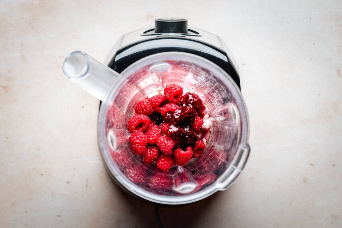 A top-down view of a blender containing fresh raspberries and raspberry preserves, placed on a light-colored countertop.