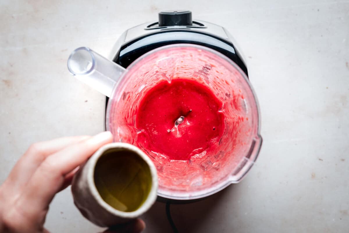 A hand holding a small cup of olive oil above a blender filled with blended raspberries, viewed from above on a light countertop.