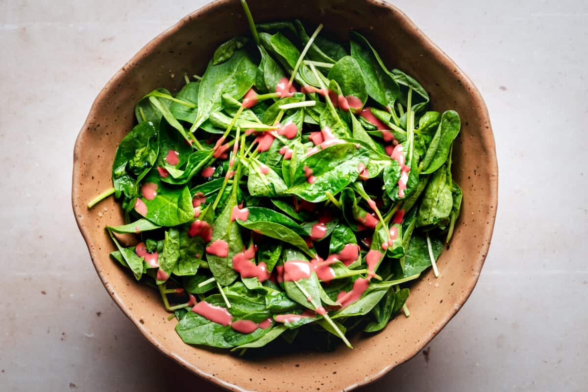 A brown bowl filled with fresh spinach leaves and topped with a drizzle of pink salad dressing, placed on a light-colored surface.