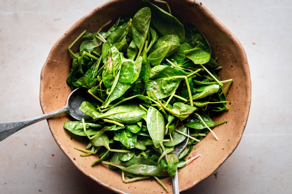A rustic brown bowl filled with fresh spinach leaves, lightly dressed, with two metal serving spoons resting inside on a light-colored surface.
