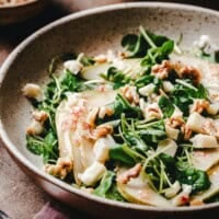 A bowl of watercress salad topped with pear slices, walnuts, and crumbled cheese sits on a table. The image has the text "Watercress Salad" and "pinchandswirl.com" at the top and bottom.