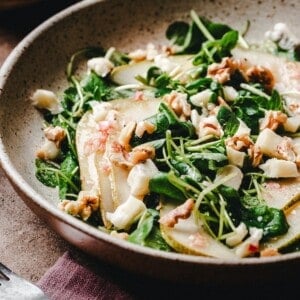 A close-up of a fresh watercress salad with sliced pears, crumbled cheese, walnuts, and microgreens in a rustic bowl on a brown surface.