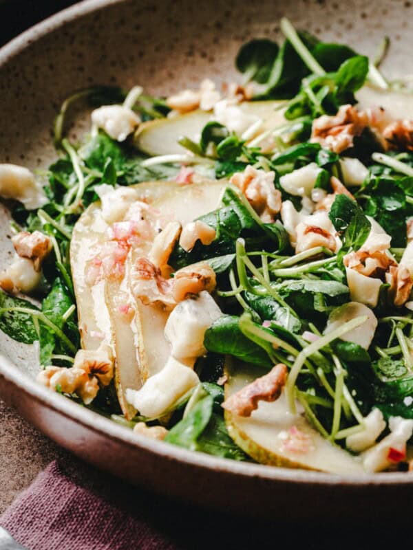 A close-up of a fresh watercress salad with sliced pears, crumbled cheese, walnuts, and microgreens in a rustic bowl on a brown surface.