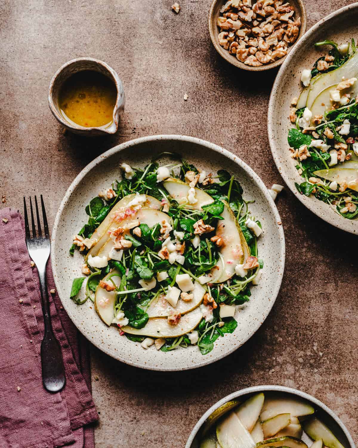 A plate of fresh watercress salad with sliced pears, crumbled gorgonzola cheese, and walnuts. A bowl of walnuts and a small jug of dressing are nearby, along with a fork and knife on a mauve cloth napkin.