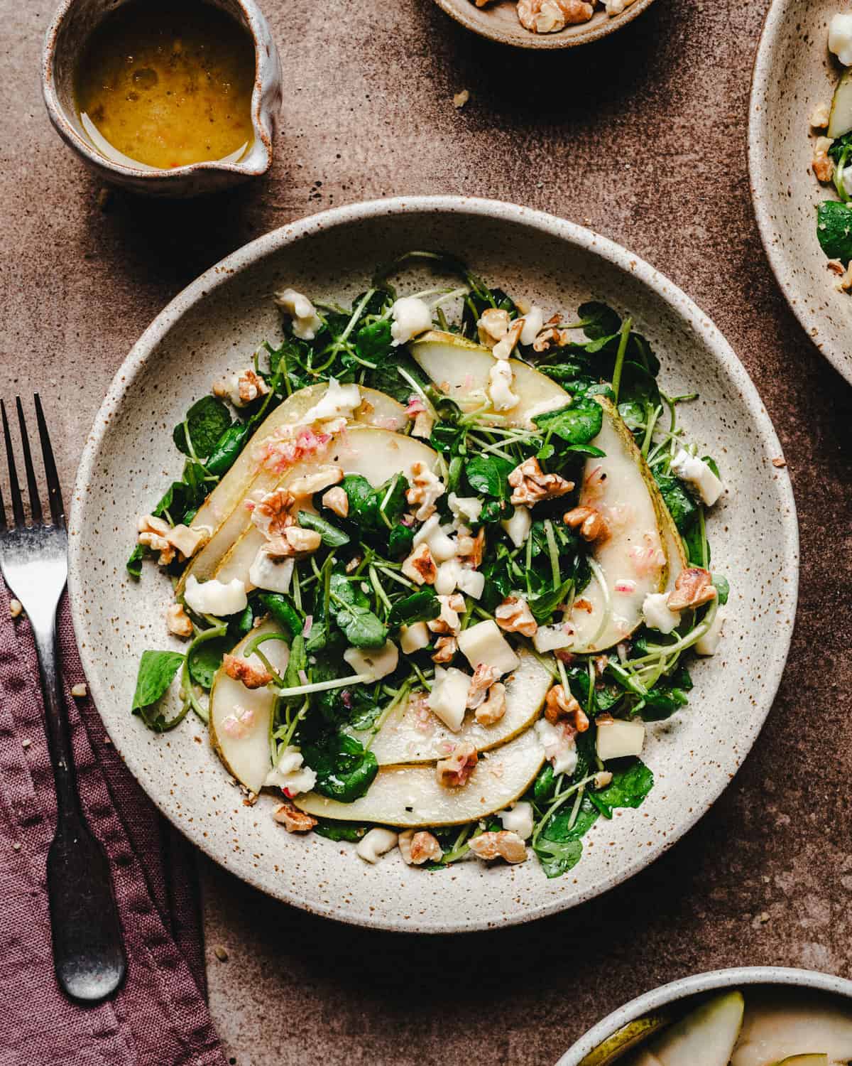 A plate of salad with watercress, pear slices, crumbled cheese, and walnuts, next to a fork on a mauve napkin and a small bowl of vinaigrette dressing on a rustic surface.