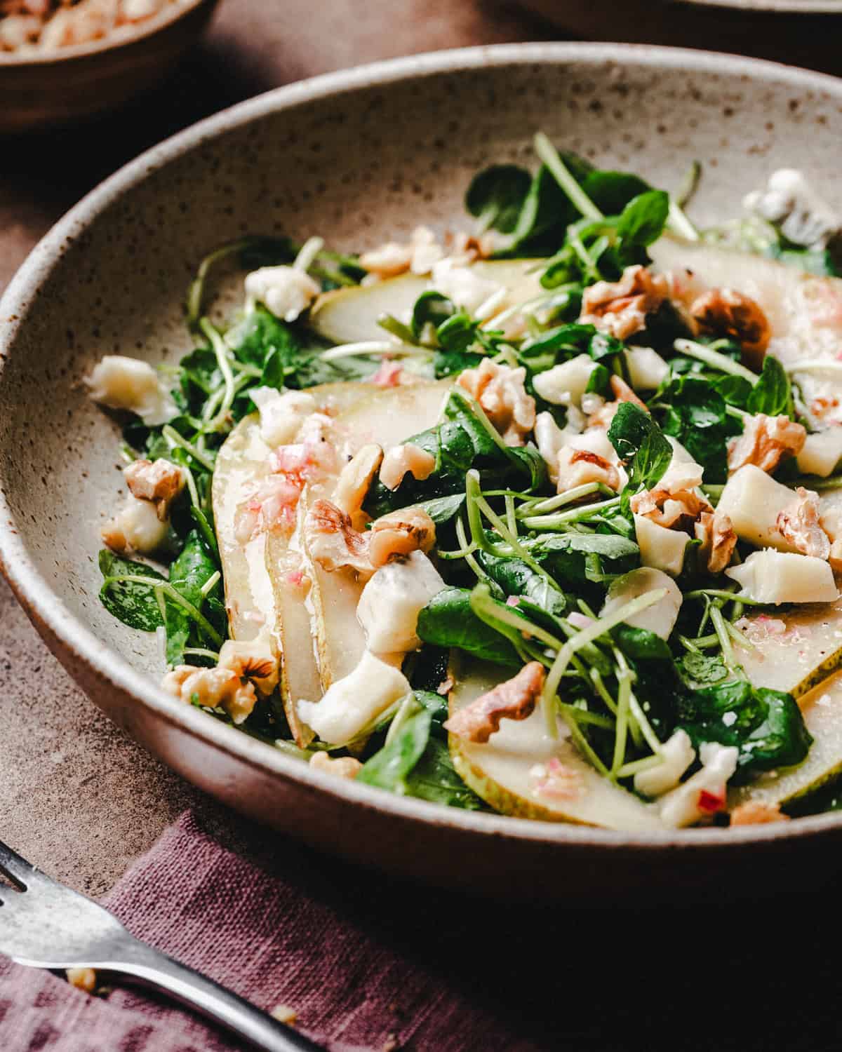 A bowl of fresh watercress salad with leafy greens, sliced pears, crumbled cheese, and walnut pieces, served on a rustic plate with a fork and napkin beside it.
