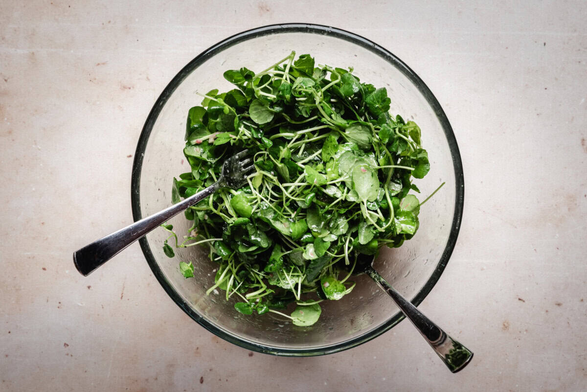 A glass bowl filled with fresh watercress, with two metal utensils resting inside, placed on a light-colored surface.