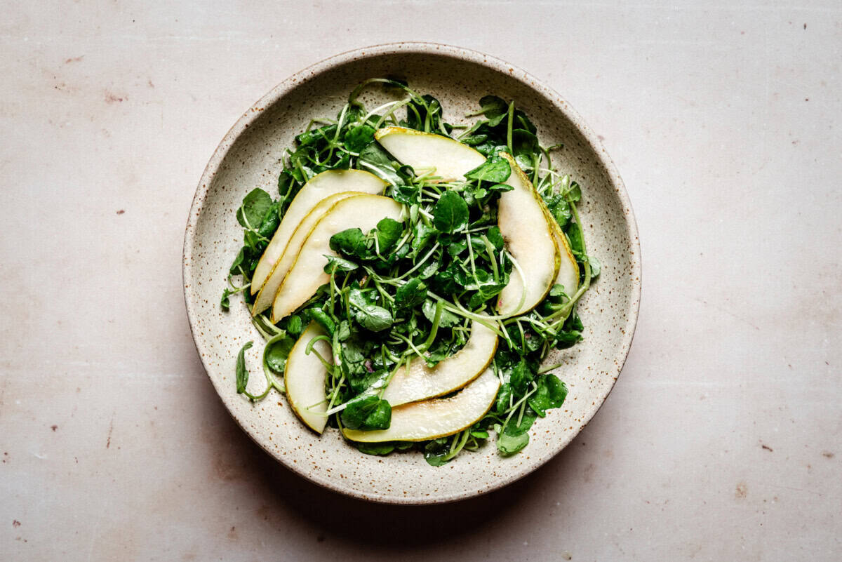 A ceramic bowl filled with a fresh salad of watercress and thinly sliced pears, arranged neatly on a light, speckled surface.