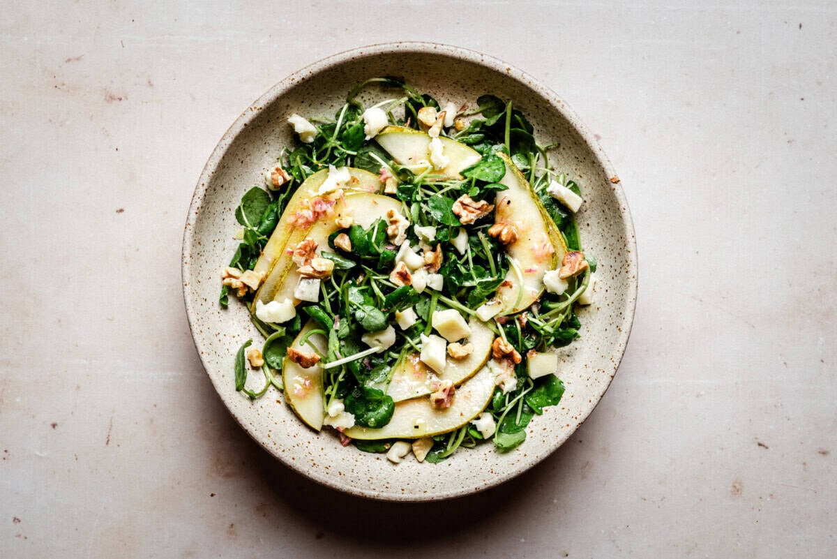 A ceramic bowl filled with a fresh salad of watercress, pear slices, walnuts, and chunks of gorgonzola cheese on a light-colored surface.