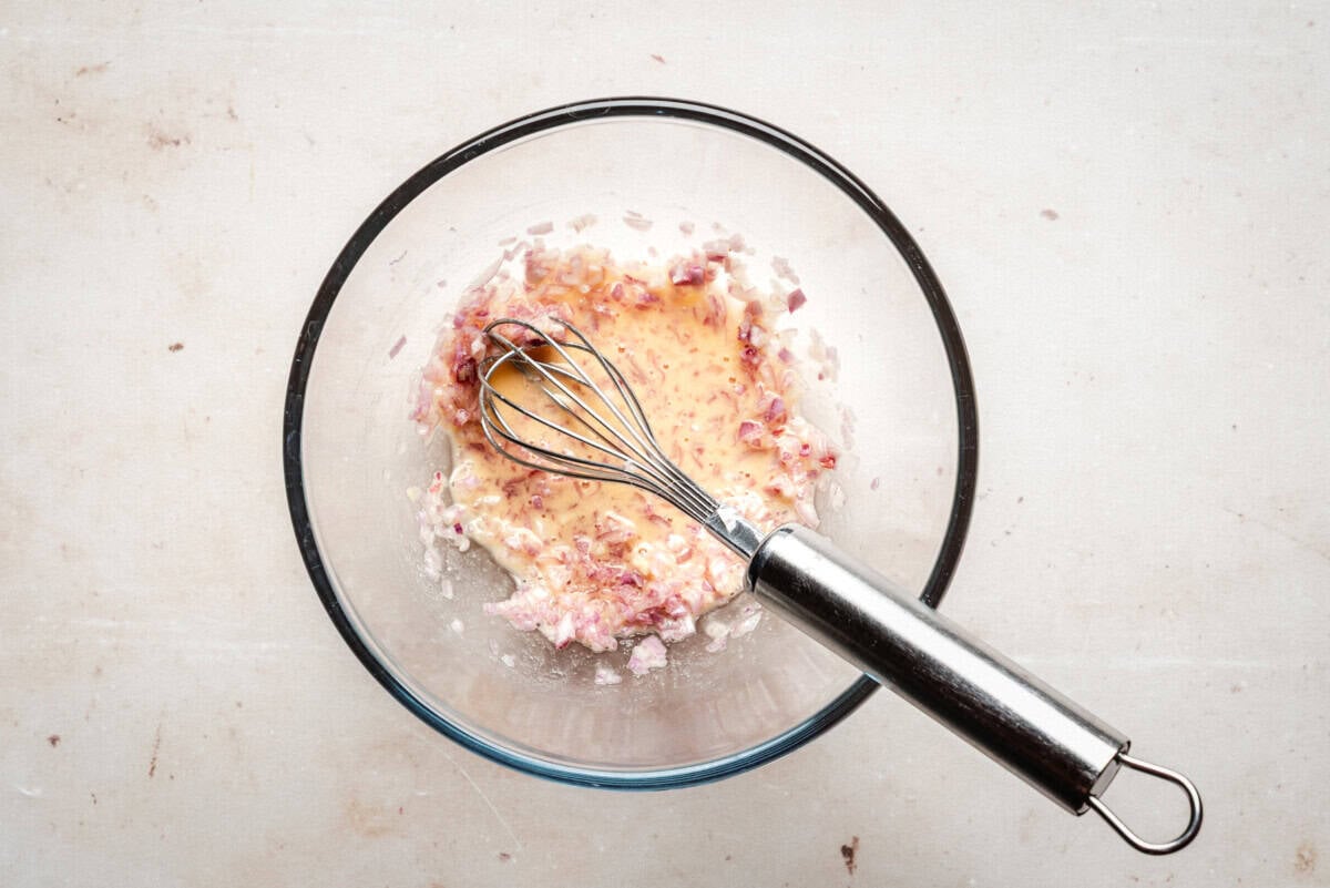A glass bowl containing a whisk and a mixture of finely chopped shallots and liquid ingredients sits on a light-colored countertop.