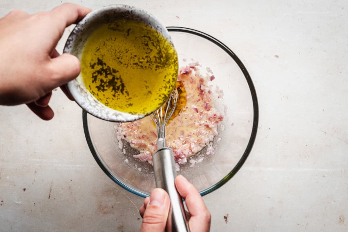 A person pours olive oil from a small bowl into a glass mixing bowl containing chopped shallot and other ingredients, while whisking the mixture with a metal whisk.