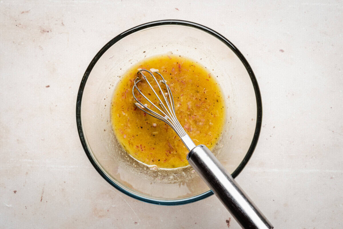 A glass bowl containing a yellowish vinaigrette with visible herbs, and a metal whisk resting inside, sits on a light-colored surface.