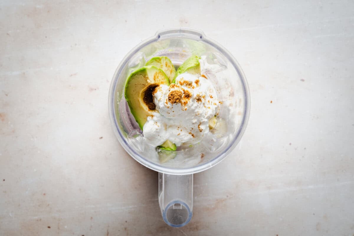 A blender cup filled with avocado halves, a dollop of sour cream, and sprinkled spices, viewed from above on a light countertop.