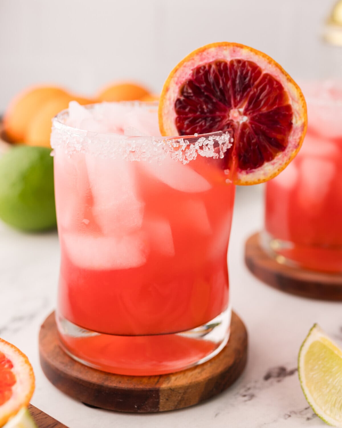 A glass of blood orange margarita cocktail with ice, garnished with a slice of blood orange and a salted rim, sits on a wooden coaster. Lime wedges and blurred fruits are in the background on a marble surface.