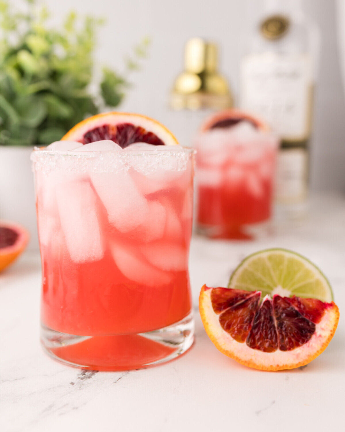 A glass of a blood orange margarita cocktail with ice, garnished with a slice of blood orange. Another similar drink, a bottle, shaker, lime, and blood orange wedge are in the blurred background on a white surface.