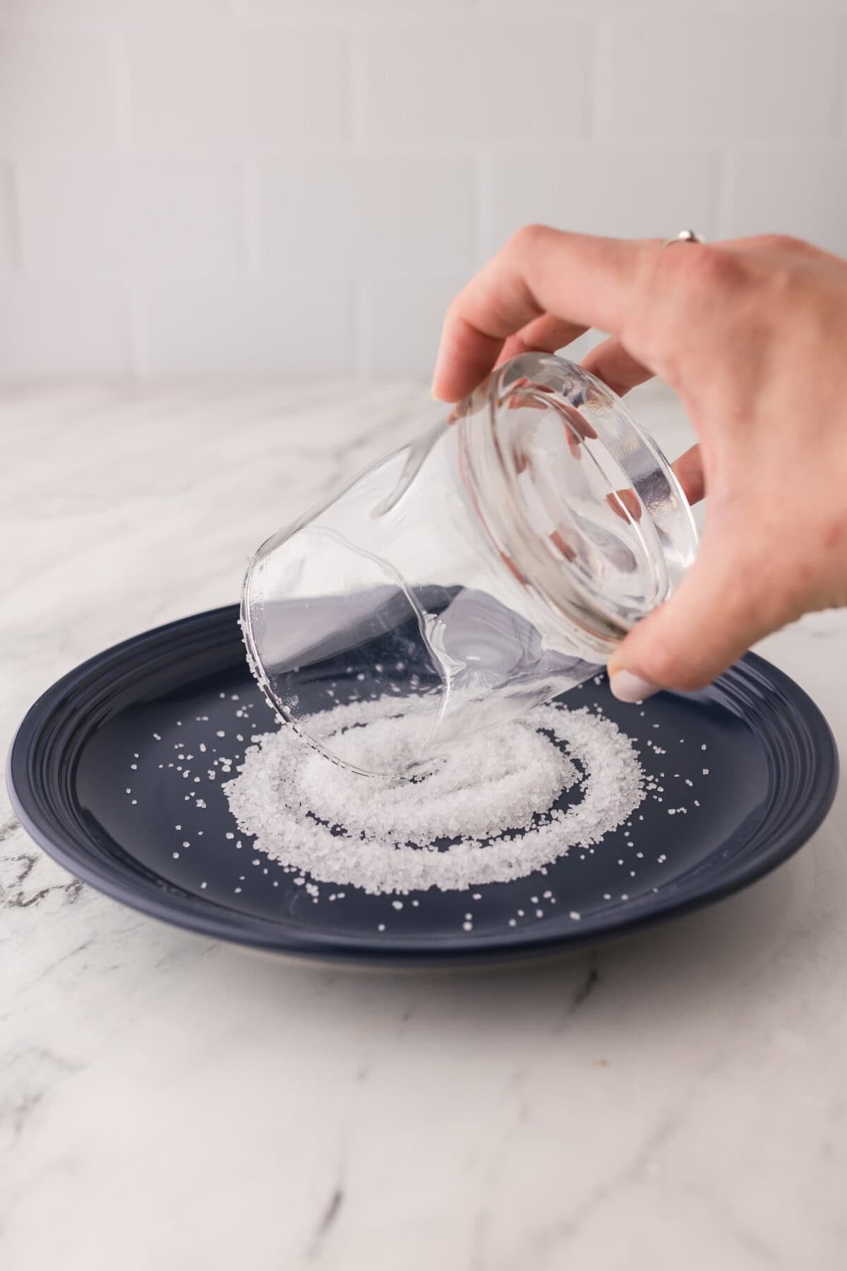 A hand is rolling the rim of a glass on a plate of salt, coating the edge of the glass. The plate is dark blue, and the surface is a light marble.
