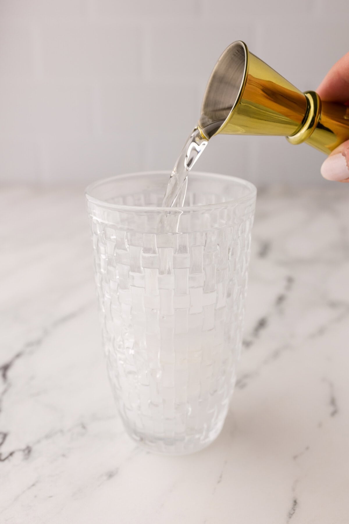 A hand pours tequila from a gold jigger into a textured glass on a white marble surface.