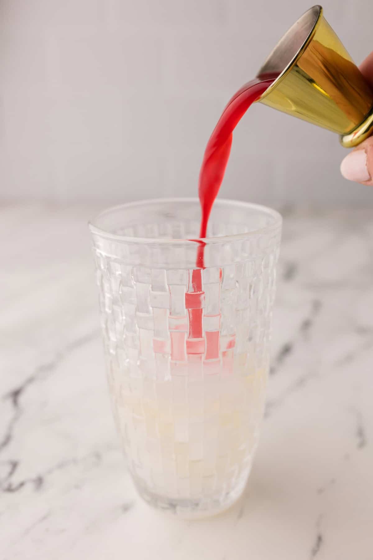 A hand pours blood orange juice from a small gold jigger into a textured glass containing a pale, cloudy liquid on a white marble surface.