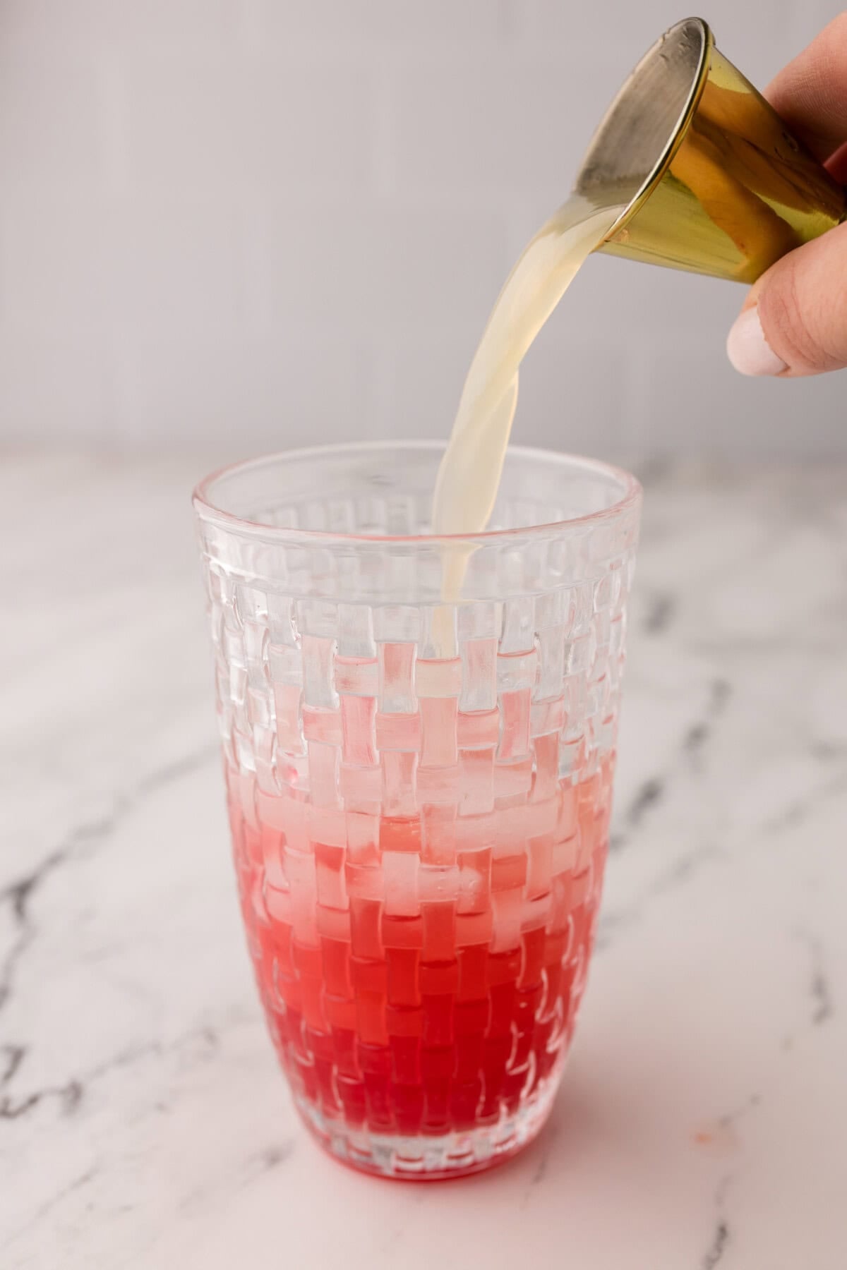 A hand pours lime juice from a jigger into a textured glass containing a red beverage with ice, set on a marbled surface.
