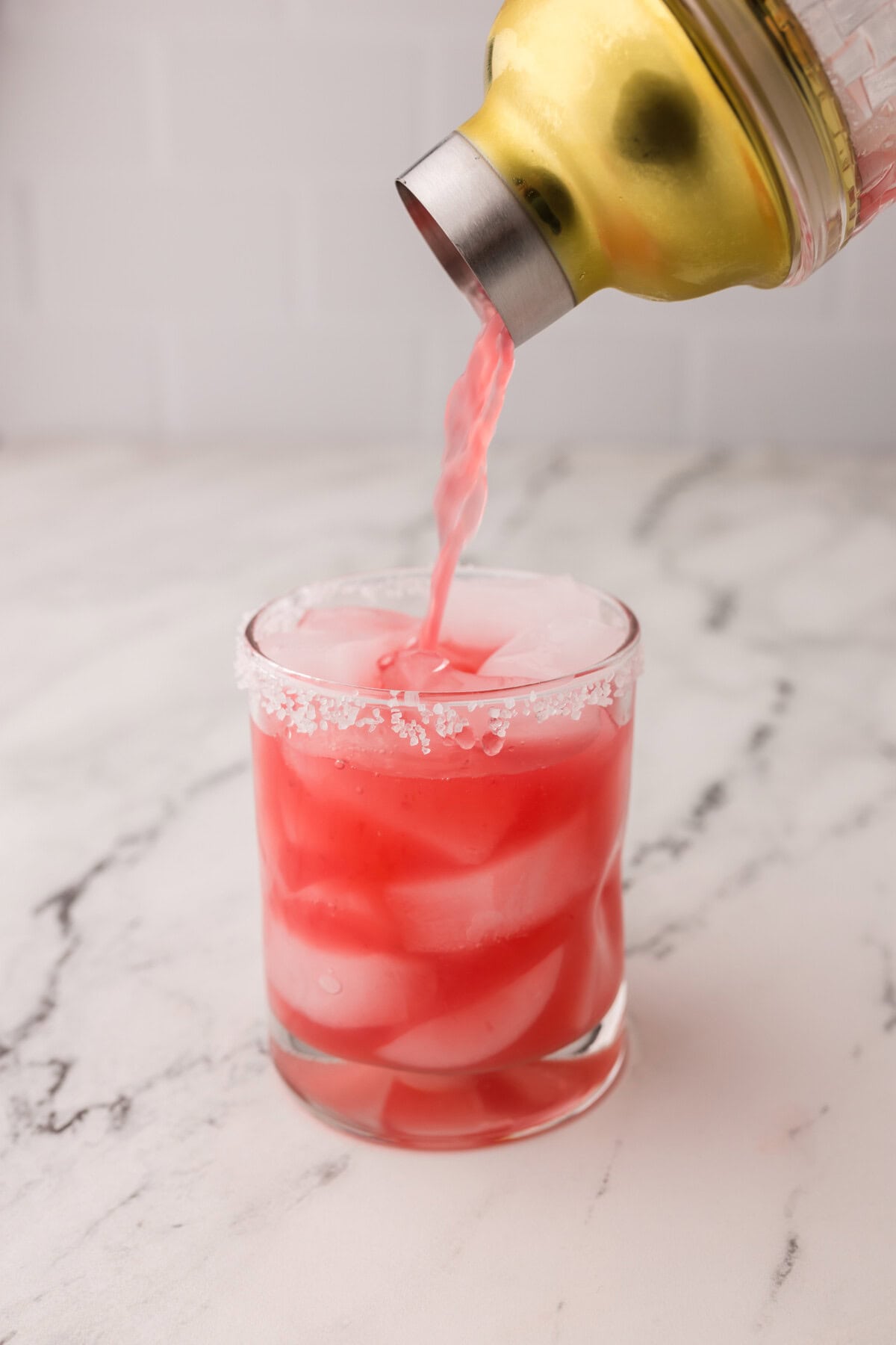 A pink blood orange margarita cocktail is being poured from a gold shaker into a glass filled with ice cubes and rimmed with salt, set on a white marble surface.