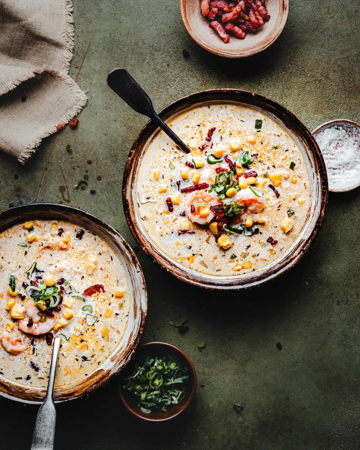 Two bowls of creamy shrimp and corn bisque topped with shrimp, corn, bacon, and herbs are on a rustic green surface. Spoons are in each bowl, with small dishes of bacon bits and sliced green onions nearby. A napkin is in the corner.
