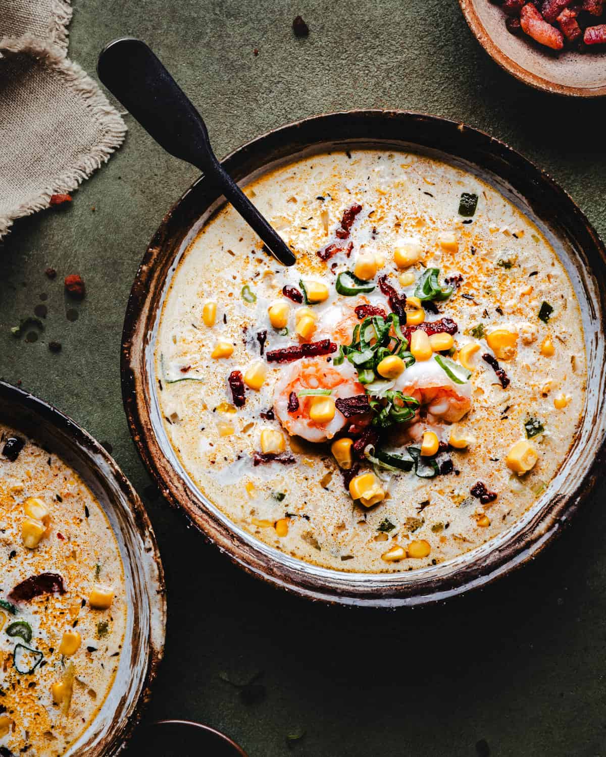 A bowl of creamy shrimp and corn bisque garnished with corn, shrimp, chopped green onions, bacon bits, and spices, with a spoon resting inside. Another partially visible bowl sits nearby on a textured surface.