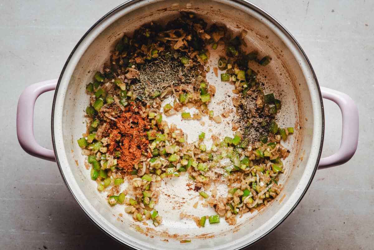 A large white pot with pink handles contains sautéed diced onions, celery, and various dried herbs and spices, including cajun seasoning, black pepper, and thyme, on a light-colored surface.