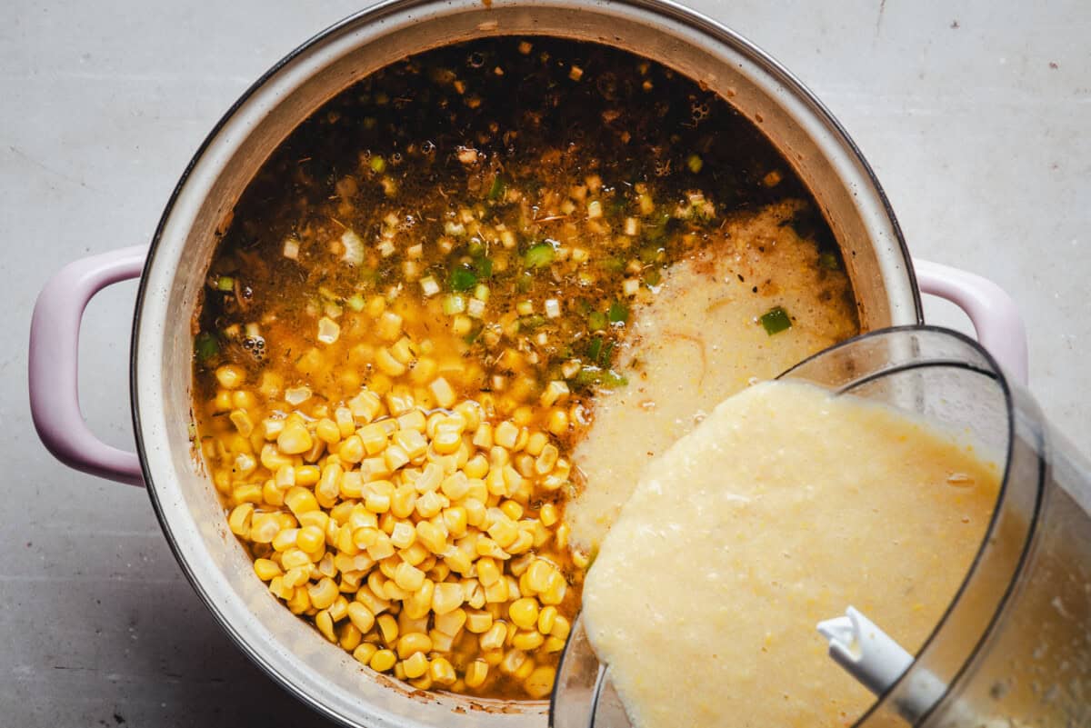 A pot filled with corn, chopped green onions, and broth sits on a countertop. Blended corn and shrimp mixture is being poured from a food processor into the pot, suggesting preparation of a corn soup or stew.