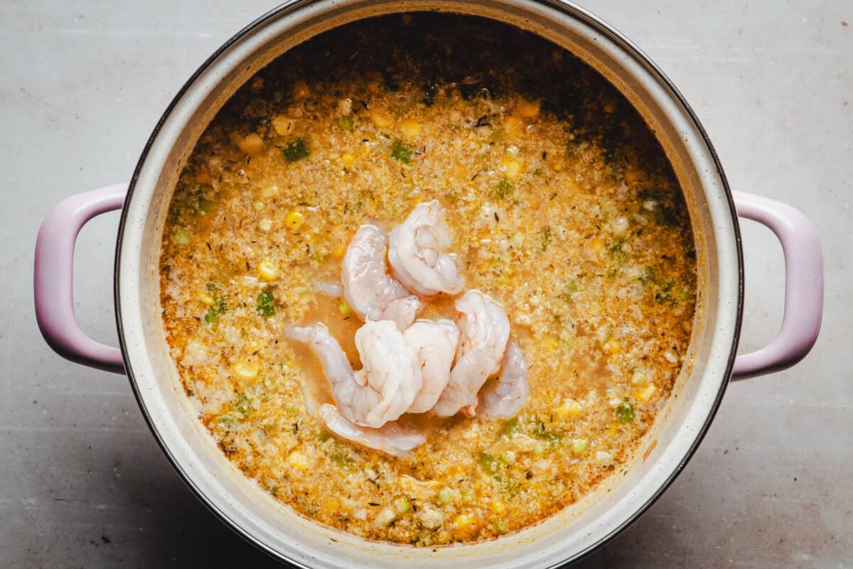 A large pot with handles contains a simmering mixture of corn, herbs, and broth, topped with several raw shrimp placed in the center.
