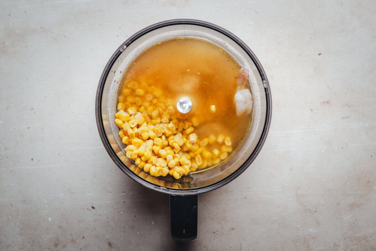 A top-down view of a food processor bowl containing corn kernels, shrimp, and broth, ready to be blended.