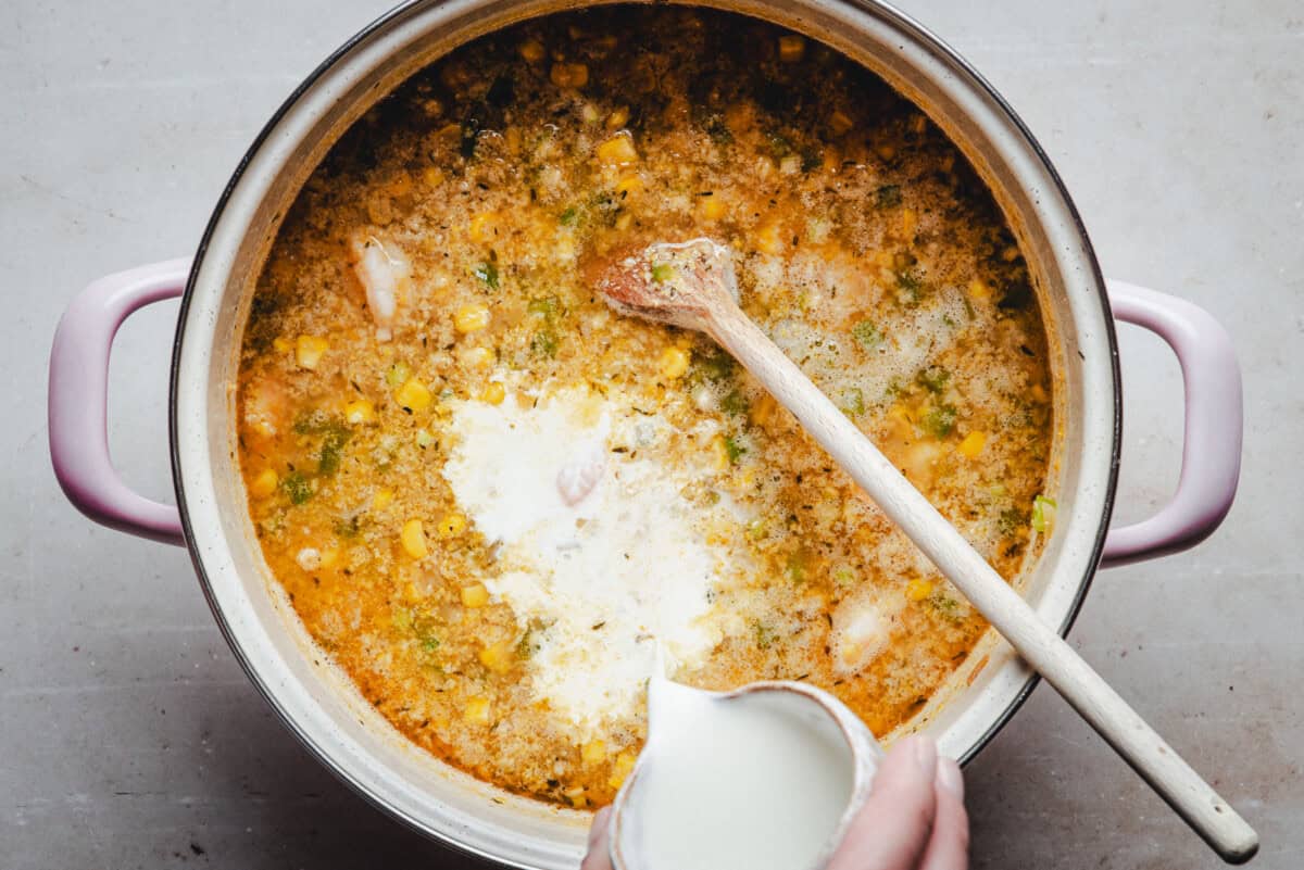 A pot of creamy bisque with corn, vegetables, and chunks of shrimp being stirred with a wooden spoon as someone adds cream from a small container.